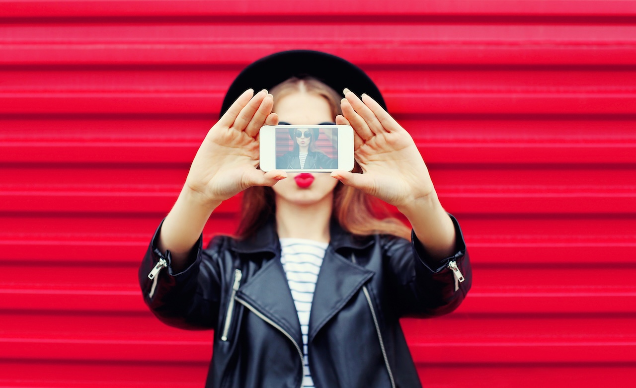 Fashion glamour woman makes self portrait on smartphone blowing lips over city pink background