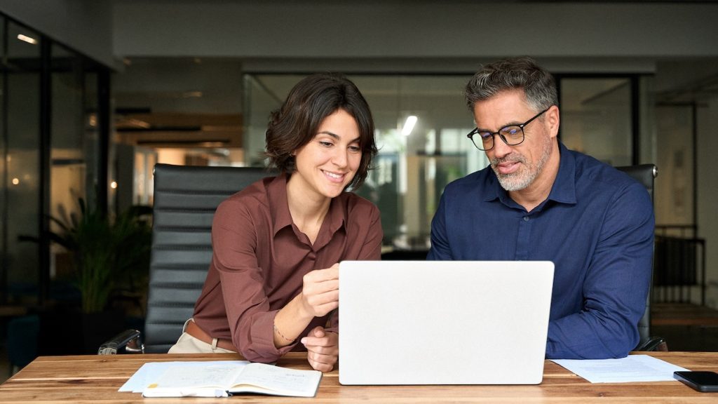 Female executive manager explaining corporate project plan to male colleague sitting at office desk. Business team of two busy professionals working together discussing ai solutions using laptop.